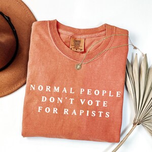 以下が含まれることがあります： 白い文字で「NORMAL PEOPLE DON'T VOTE FOR RAPISTS」と書かれた、コーラル色のComfort Colorsスウェットシャツ。ペンダント付きのゴールドネックレスがシャツの上にかけられています。茶色の帽子と乾燥した植物が背景にあります。