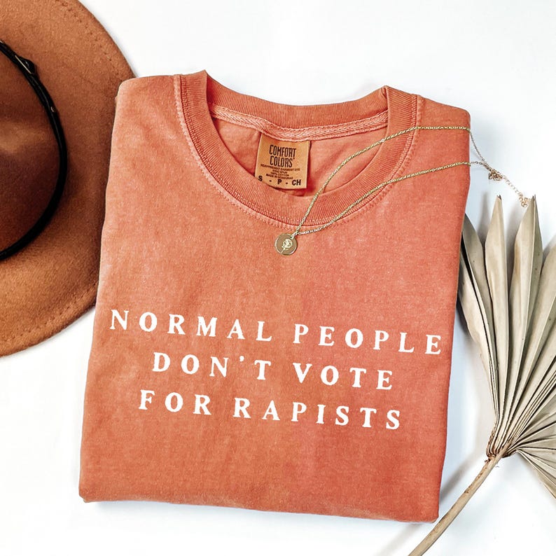 以下が含まれることがあります： 白い文字で「NORMAL PEOPLE DON'T VOTE FOR RAPISTS」と書かれた、コーラル色のComfort Colorsスウェットシャツ。ペンダント付きのゴールドネックレスがシャツの上にかけられています。茶色の帽子と乾燥した植物が背景にあります。