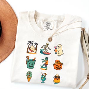 May include: Cream-colored Comfort Colors t-shirt with colorful Halloween-themed graphics. The shirt features illustrations of a ghost in a hammock, a skeleton surfing, a ghost with a drink, a jack-o'-lantern bucket, a zombie, and a pumpkin.