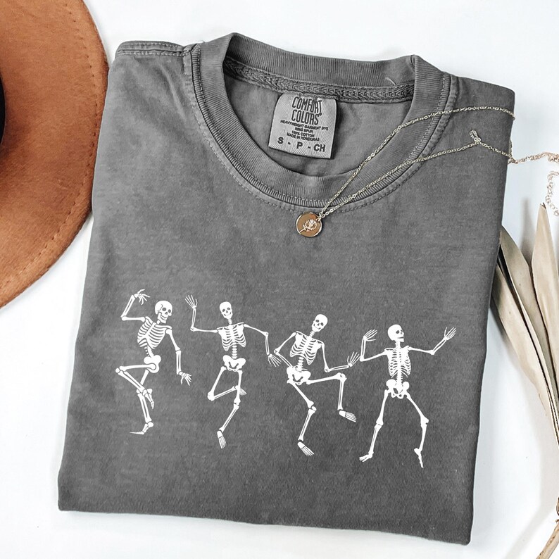May include: A grey Comfort Colors t-shirt with a white graphic of dancing skeletons. The shirt has a crew neck and short sleeves. A silver necklace with a pendant is draped across the shirt. The tag reads "S - P - CH".