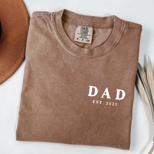 May include: A brown Comfort Colors t-shirt with the word "DAD" and "EST. 2025" printed in white. The shirt is folded and has a crew neck. A brown hat and dried leaves are in the background.