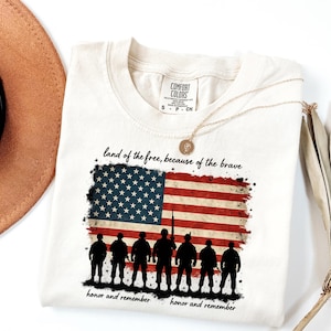 May include: A white t-shirt with a distressed American flag design and the text "land of the free, because of the brave" and "honor and remember". Silhouettes of soldiers are standing in front of the flag.