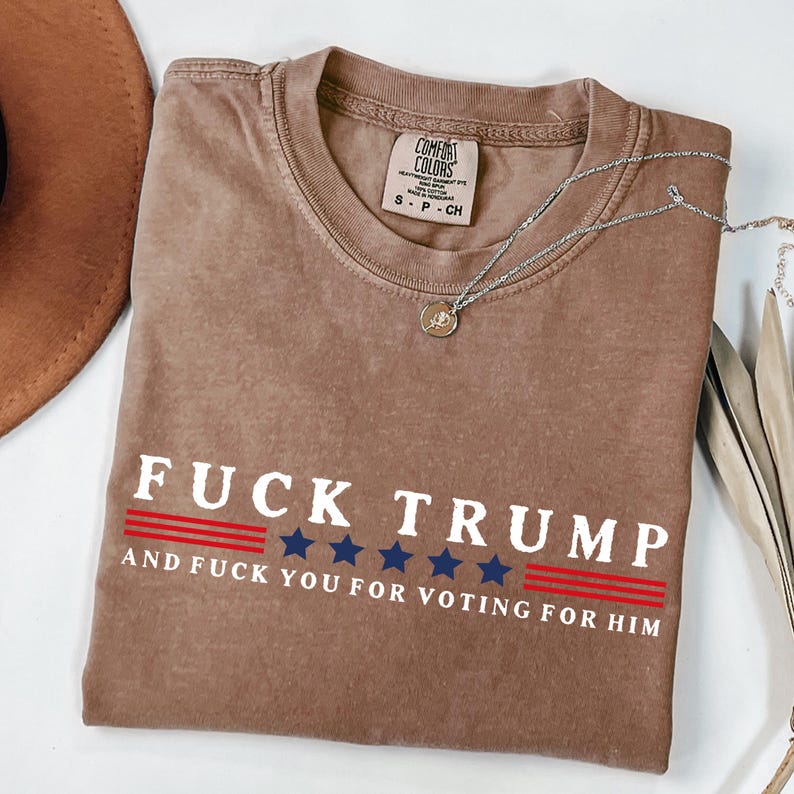 May include: A brown Comfort Colors t-shirt with the text "FUCK TRUMP" in white, with red and blue accents. The shirt also includes the phrase "AND FUCK YOU FOR VOTING FOR HIM". A silver necklace with a pendant is draped across the shirt.