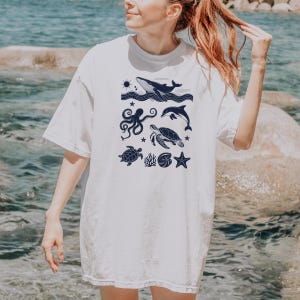 May include: White t-shirt featuring a navy blue illustration of marine life. The design includes a whale, dolphin, octopus, sea turtle, starfish, and other sea creatures. The shirt is a casual, oversized fit.
