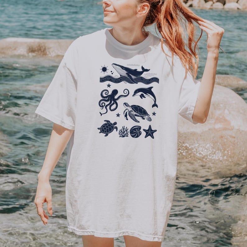May include: White t-shirt featuring a navy blue illustration of marine life. The design includes a whale, dolphin, octopus, sea turtle, starfish, and other sea creatures. The shirt is a casual, oversized fit.