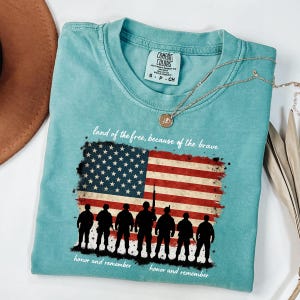May include: A teal blue t-shirt with a distressed American flag design and the text "land of the free, because of the brave" and "honor and remember". Silhouettes of soldiers are standing in front of the flag.