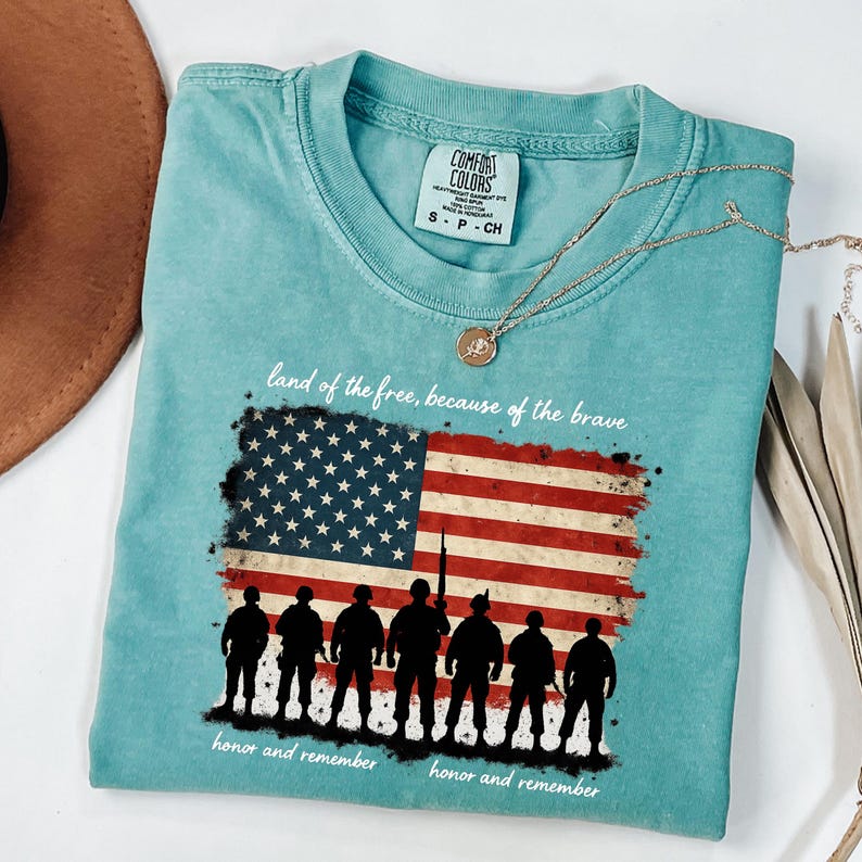 May include: A teal blue t-shirt with a distressed American flag design and the text "land of the free, because of the brave" and "honor and remember". Silhouettes of soldiers are standing in front of the flag.