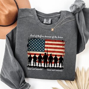 May include: A dark gray sweatshirt with a distressed American flag design and the text "land of the free, because of the brave".  Below the flag are seven silhouettes of soldiers with the text "honor and remember" on either side.