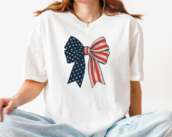 May include: White t-shirt with a red, white, and blue bow printed on the front. The bow is designed to look like an American flag.