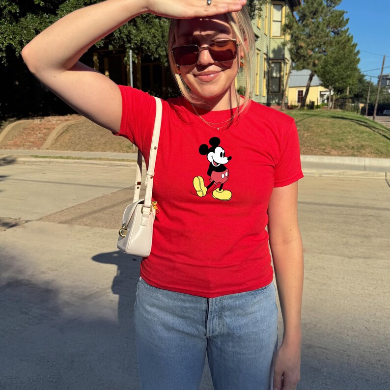 May include: A red t-shirt featuring a classic Mickey Mouse graphic. The shirt is paired with light blue jeans and a small, white purse. The person is wearing sunglasses and standing outside on a sunny day.