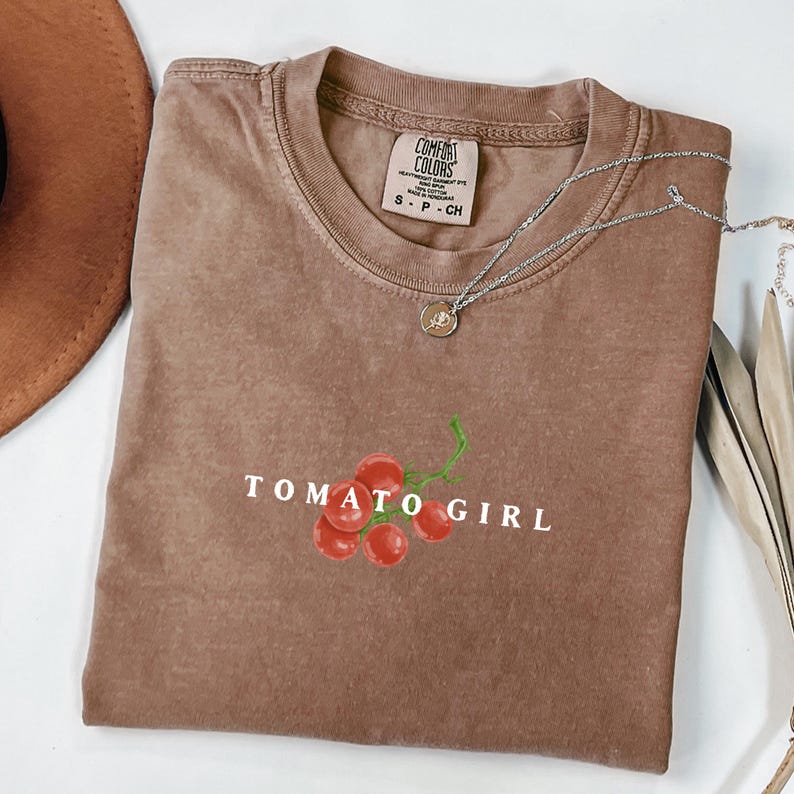 May include: A brown t-shirt with a white graphic of a bunch of red tomatoes with a green stem and the text "TOMATO GIRL".