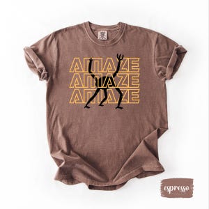 May include: Espresso-colored t-shirt with the word "AMAZE" stacked three times in yellow, with a black figure in the center. The shirt is made of a soft material and has a vintage look.