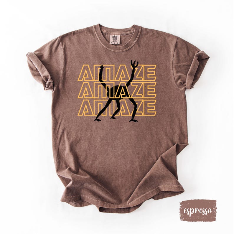 May include: Espresso-colored t-shirt with the word "AMAZE" stacked three times in yellow, with a black figure in the center. The shirt is made of a soft material and has a vintage look.