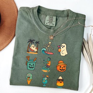 May include: Olive green t-shirt with a variety of Halloween-themed cartoon graphics. The graphics include a ghost in a hammock, a skeleton surfing, a ghost with a cocktail, a jack-o'-lantern bucket, a zombie, and a jack-o'-lantern wearing a hat.