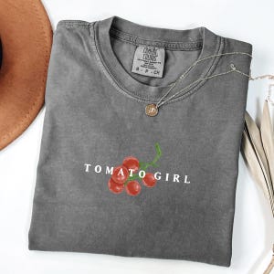 May include: Gray t-shirt with a white graphic of a cluster of red tomatoes with a green stem and the text "TOMATO GIRL".