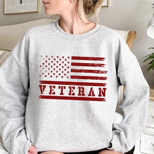 May include: Gray sweatshirt with a red, white, and blue distressed American flag and the word "VETERAN" in red.