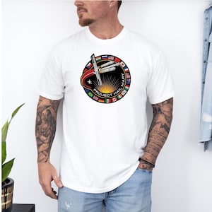 May include: White t-shirt featuring a circular graphic with the text "PROJECT HAIL MARY". The graphic includes a stylized rocket, a sunburst design, and flags around the perimeter. The shirt is short-sleeved.