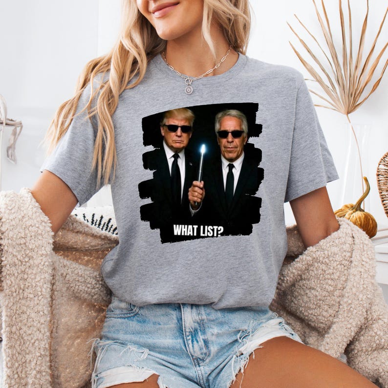 K&ouml;nnte beinhalten: Ein graues T-Shirt mit einer Grafik von zwei M&auml;nnern in schwarzen Anz&uuml;gen und Sonnenbrillen, die einen leuchtenden Gegenstand halten. Der Text "WHAT LIST?" steht unter dem Bild. Das T-Shirt ist ein Freizeitkleidungsst&uuml;ck.