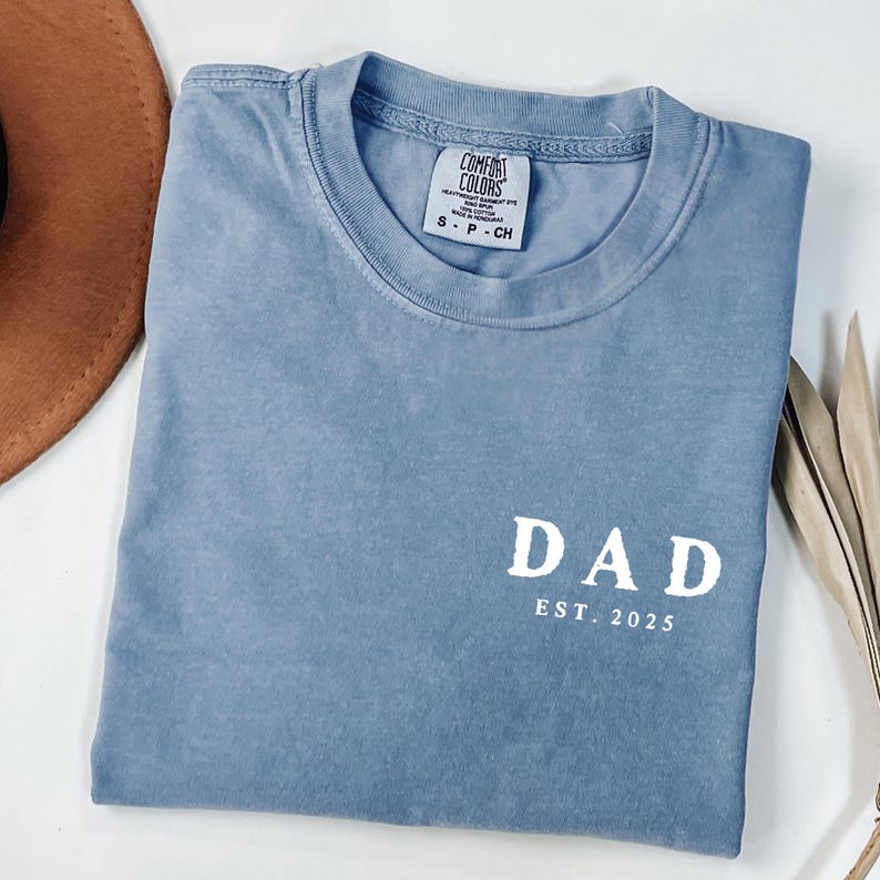May include: A blue Comfort Colors t-shirt with the word "DAD" and "EST. 2025" printed in white. The shirt is folded and has a small tag on the collar. A brown hat and dried plant are in the background.