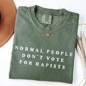 以下が含まれることがあります： 「NORMAL PEOPLE DON'T VOTE FOR RAPISTS」の白文字がプリントされたオリーブグリーンのTシャツ。クルーネックと半袖です。ペンダント付きのゴールドネックレスがシャツの上にかけられています。