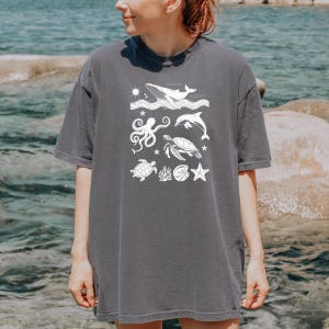 May include: A charcoal gray t-shirt featuring a white graphic of various sea creatures. The design includes a whale, dolphin, octopus, sea turtle, starfish, and other marine elements. The shirt has short sleeves and a relaxed fit.