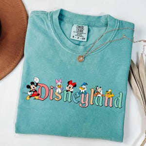 May include: A turquoise Comfort Colors t-shirt featuring the word "Disneyland" in colorful lettering, with Mickey Mouse and friends. A gold necklace and a brown hat are also visible.