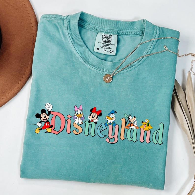May include: A turquoise Comfort Colors t-shirt featuring the word "Disneyland" in colorful lettering, with Mickey Mouse and friends. A gold necklace and a brown hat are also visible.