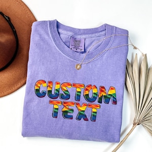 May include: A lavender Comfort Colors t-shirt with the words "CUSTOM TEXT" in rainbow-colored bubble letters. A gold necklace with a pendant is draped over the shirt. A brown hat and dried plant are in the background.