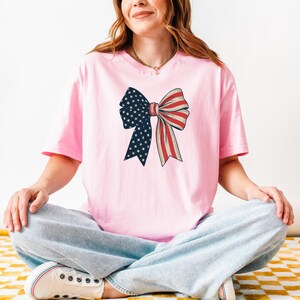 Peut inclure: Un t-shirt rose avec un grand n&oelig;ud imprim&eacute; sur le devant. Le n&oelig;ud est aux couleurs du drapeau am&eacute;ricain, rouge, blanc et bleu.