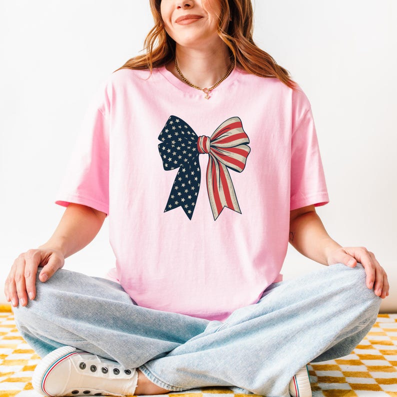 Peut inclure: Un t-shirt rose avec un grand n&oelig;ud imprim&eacute; sur le devant. Le n&oelig;ud est aux couleurs du drapeau am&eacute;ricain, rouge, blanc et bleu.