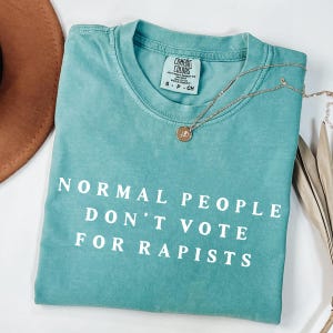 以下が含まれることがあります： 「NORMAL PEOPLE DON'T VOTE FOR RAPISTS」の白文字がプリントされた、ターコイズ色のComfort Colors Tシャツ。 金色のネックレスがシャツの上に置かれています。 茶色の帽子と乾燥した葉が背景にあります。