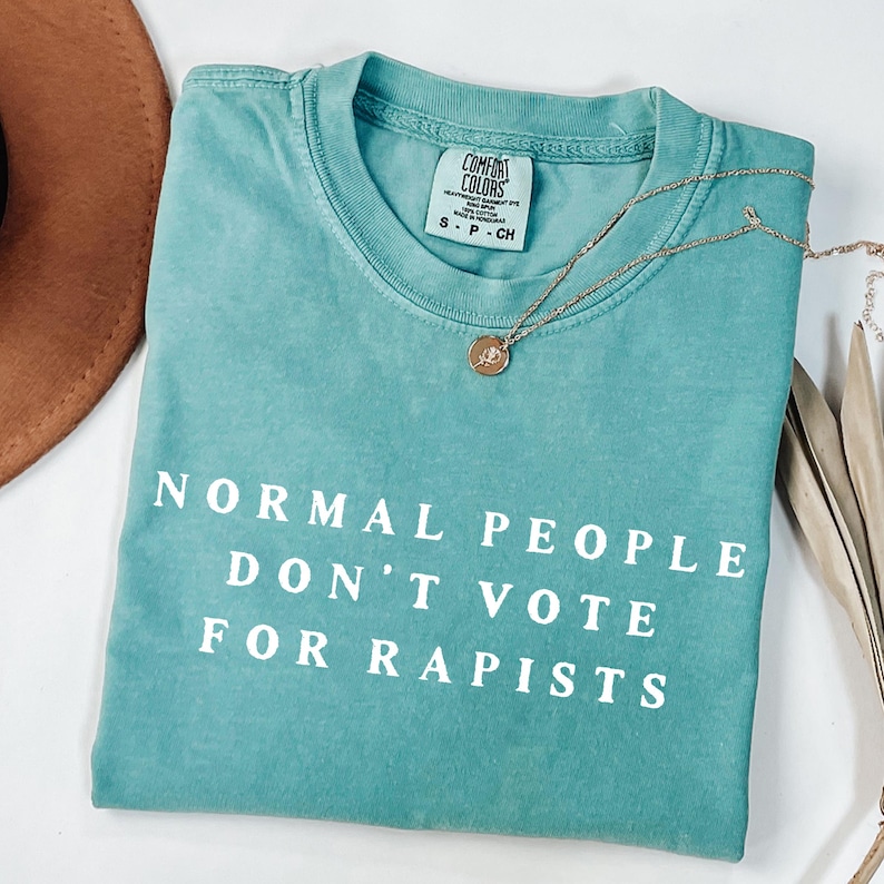 以下が含まれることがあります： 「NORMAL PEOPLE DON'T VOTE FOR RAPISTS」の白文字がプリントされた、ターコイズ色のComfort Colors Tシャツ。 金色のネックレスがシャツの上に置かれています。 茶色の帽子と乾燥した葉が背景にあります。
