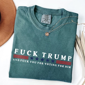 May include: A teal Comfort Colors t-shirt with the text "FUCK TRUMP" in white block letters, with red and blue accents. The shirt also includes the phrase "AND FUCK YOU FOR VOTING FOR HIM". A gold necklace is draped across the shirt.