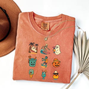 May include: Coral-colored Comfort Colors sweatshirt featuring a grid of cartoon graphics. Designs include a ghost in a hammock, a skeleton surfing, a ghost with a drink, a bucket, a zombie, a jack-o'-lantern, ice cream, a cocktail, and a candy corn.