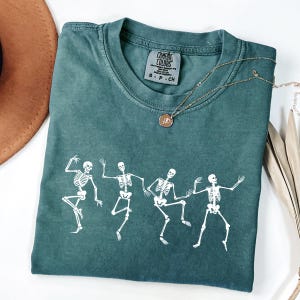 May include: Teal Comfort Colors t-shirt with a white graphic of dancing skeletons. The shirt has a crew neck and short sleeves. A gold necklace with a pendant is draped across the shirt. The shirt is folded.