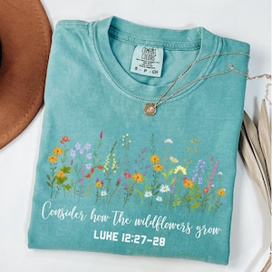 May include: A teal blue t-shirt with a floral design and the text "Consider how the wildflowers grow. LUKE 12:27-28"