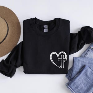 May include: Black sweatshirt with a white heart and cross design. The heart is outlined and the cross is inside the heart.