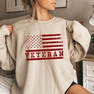 May include: A beige sweatshirt with a red, white, and blue American flag graphic and the word "VETERAN" printed in red.