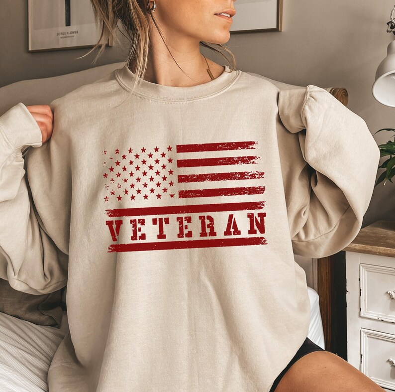 May include: A beige sweatshirt with a red, white, and blue American flag graphic and the word "VETERAN" printed in red.