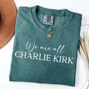 May include: A teal Comfort Colors t-shirt with the text "We are all CHARLIE KIRK" in white script. A gold necklace with a pendant rests on the shirt. A brown hat and dried plant stems are in the background.