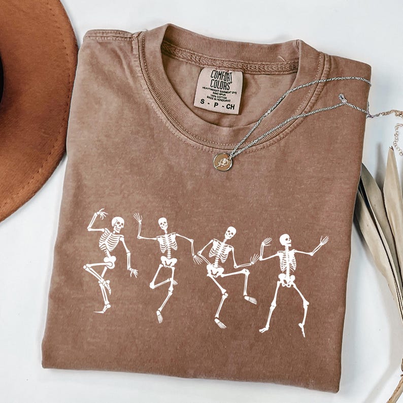 May include: A brown Comfort Colors t-shirt featuring a white graphic of dancing skeletons. The t-shirt has a crew neck and short sleeves. A silver necklace with a pendant is draped across the shirt. The t-shirt is size small.