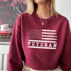 May include: A maroon sweatshirt with a white distressed American flag and the word "VETERAN" printed on the front.
