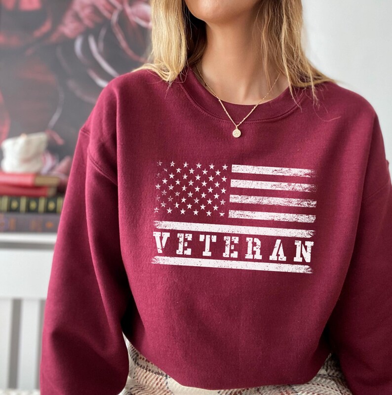 May include: A maroon sweatshirt with a white distressed American flag and the word "VETERAN" printed on the front.