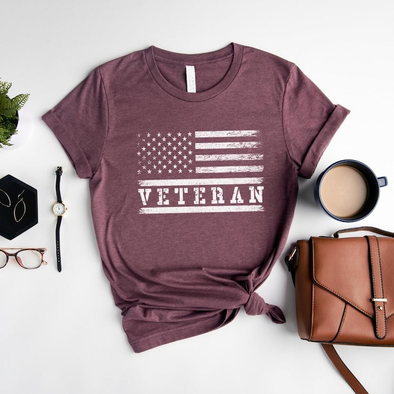 以下が含まれることがあります： 白くぼやけたアメリカの旗と「Veteran」という文字がプリントされた、マルーンのTシャツです。