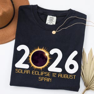 May include: A black t-shirt featuring the white text 2026 with a graphic of a solar eclipse replacing the zero. Below the graphic, the text reads SOLAR ECLIPSE 12 AUGUST SPAIN in yellow. The shirt has a Comfort Colors brand label at the neck.