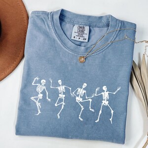 May include: A blue Comfort Colors t-shirt with a white print of five dancing skeletons. A gold necklace with a pendant lies on the shirt. The shirt is folded, with a brown hat visible in the upper left corner.