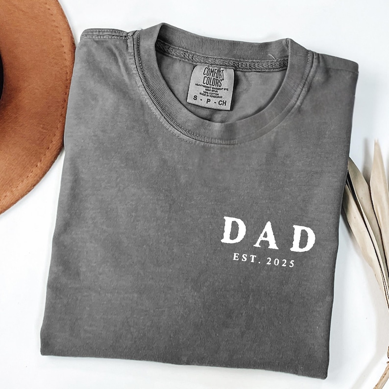 May include: A gray Comfort Colors t-shirt with the word "DAD" and "EST. 2025" printed in white. The shirt is folded and placed on a white surface, with a brown hat and dried plant stems in the background.