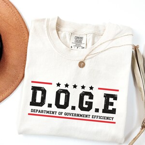 May include: A white t-shirt with a red and black graphic that says "D.O.G.E. DEPARTMENT OF GOVERNMENT EFFICIENCY".