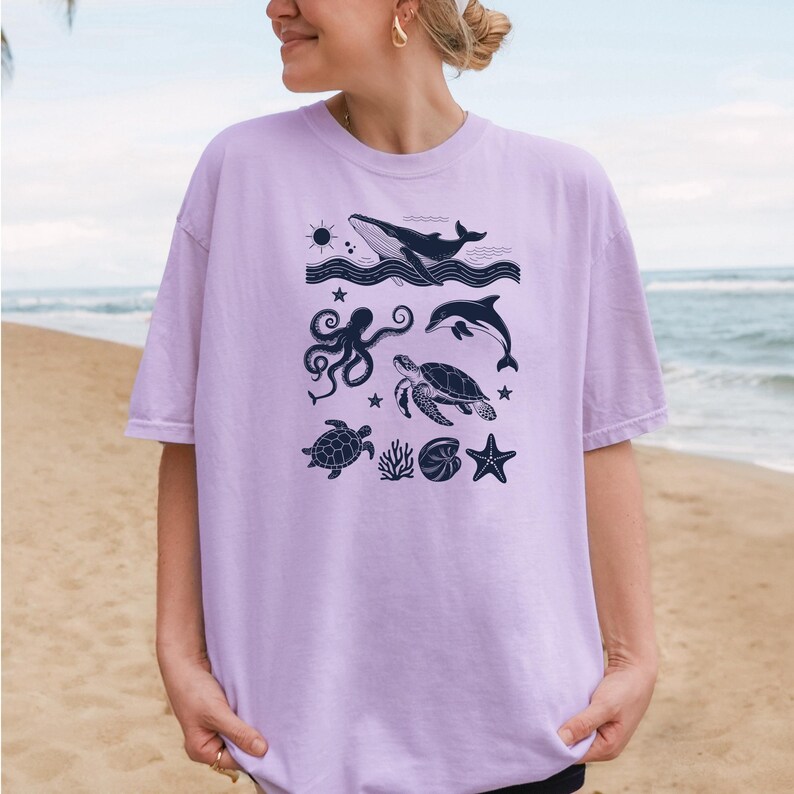 May include: A light purple t-shirt featuring a navy blue ocean-themed graphic. The design includes a whale, dolphin, octopus, sea turtle, starfish, and other sea elements. The shirt is a relaxed fit.