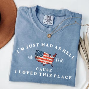 May include: A blue short-sleeved t-shirt with a Comfort Colors label. The graphic features a map of the United States filled with the American flag, the text "I'M JUST MAD AS HELL", "est. 1776", and "CAUSE I LOVED THIS PLACE" in white lettering.